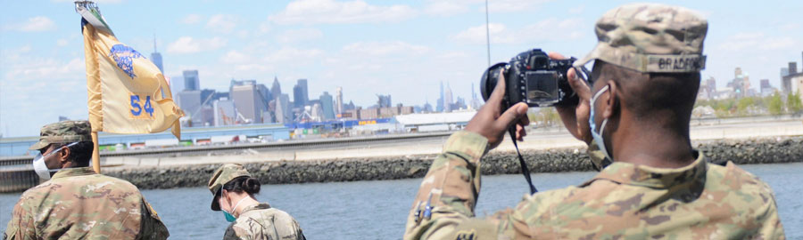  The image shows a group of military personnel wearing camouflage uniforms and face masks, standing outdoors near a body of water. One soldier, in the foreground on the right, is taking a photograph using a camera. Another soldier on the left is holding a flag with the number "54" visible on it. In the background, across the water, there is a city skyline with several tall buildings under a clear blue sky with scattered clouds. This setting appears to be near a waterfront or port area with the cityscape as a backdrop.